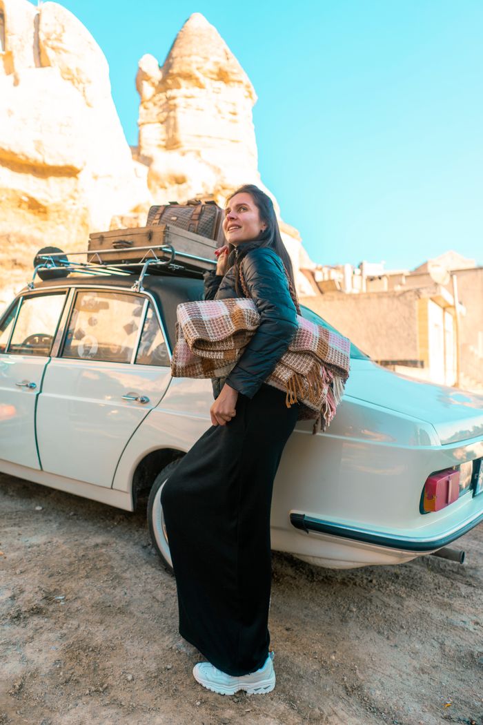 about-03 Smiling woman leaning on a vintage car with rocky formations in Cappadocia, Turkey.