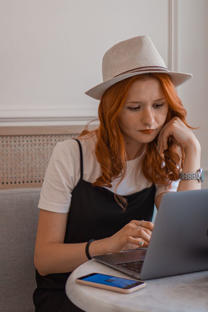 menu-10 Redhead woman wearing a hat, focused on laptop work with a smartphone on the table.