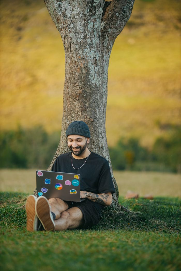 menu-08 Young man with beard sits under a tree, smiling and using a laptop in a serene meadow.