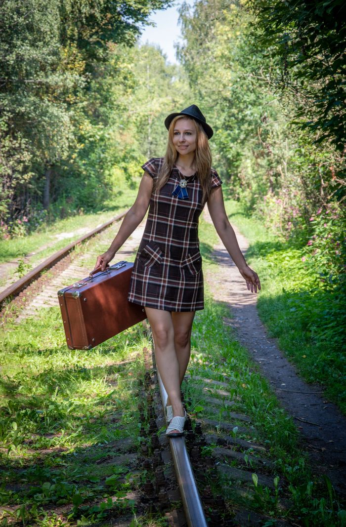about-02 A cheerful woman in a sundress balancing on a railway track with a suitcase in a lush forest setting.