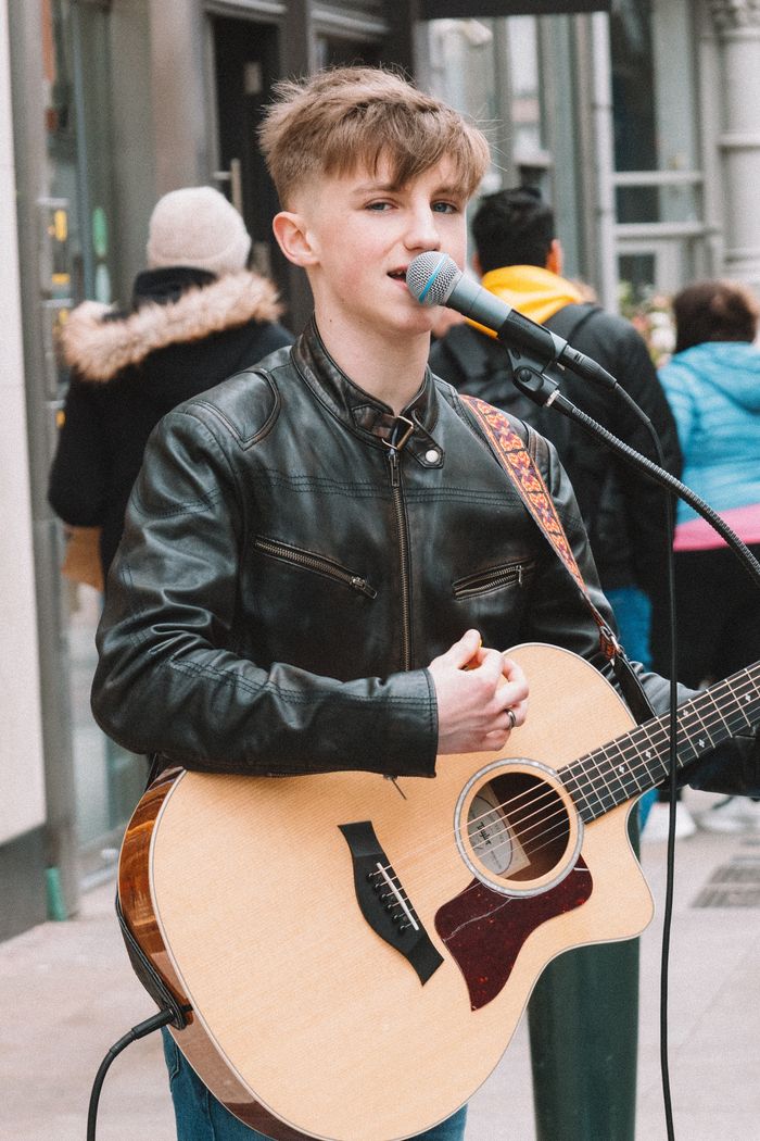menu-03 Street musician in a black leather jacket playing acoustic guitar and singing in an urban setting.
