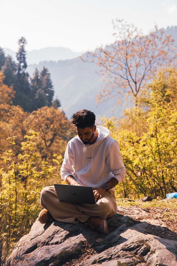 menu-09 A man sitting on a rock in a forest, using a laptop for remote work in a tranquil natural setting.