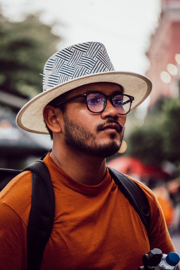 menu-18 Portrait of a young man outdoors wearing a stylish hat and glasses, with a blurred urban background.