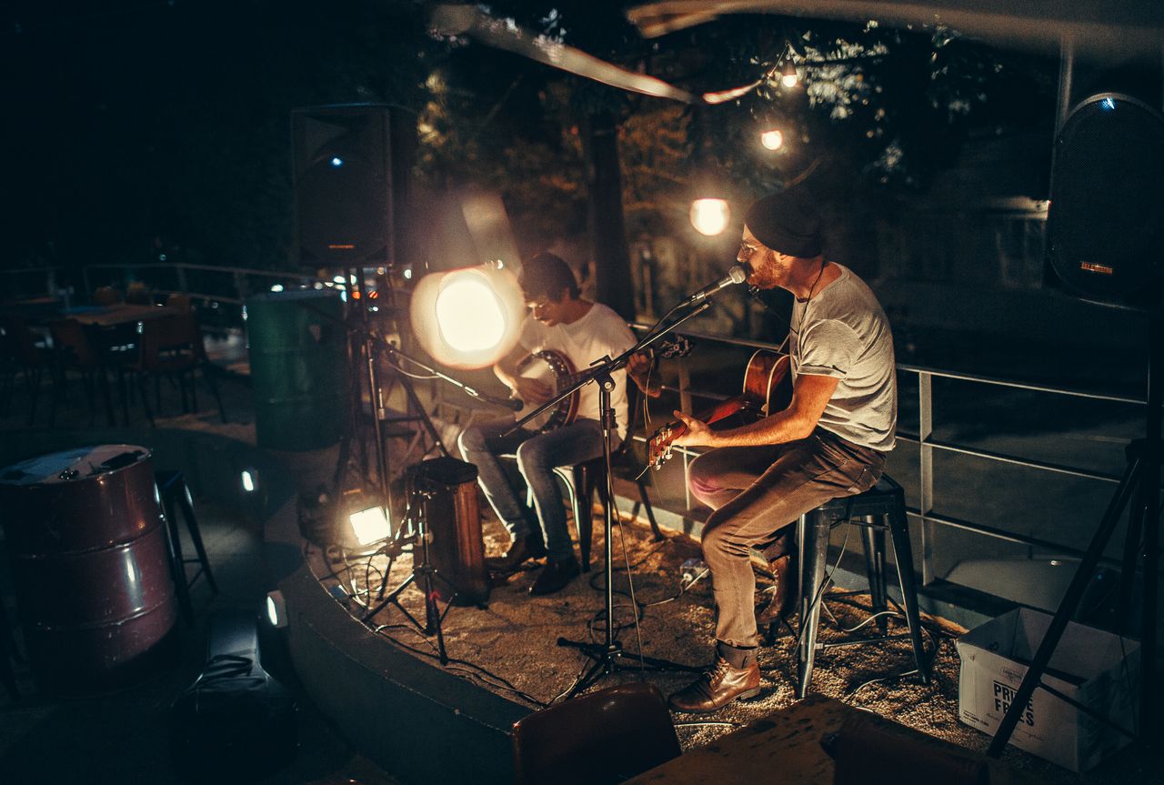 menu-01 Two musicians performing acoustic music outdoors at night, illuminated by soft ambient lighting.