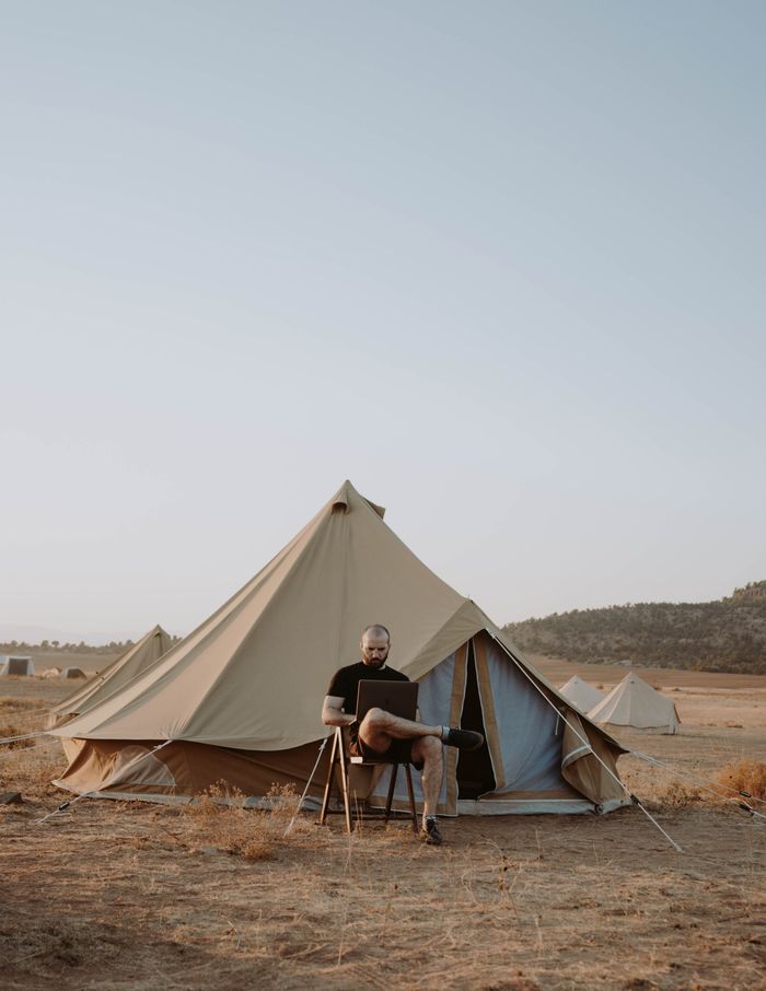 Full body of bearded male freelancer sitting on chair with laptop and working during camping in countryside