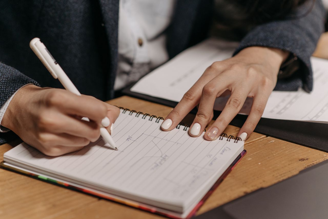 Close-up of a person writing notes in a spiral notebook with a pen on a wooden desk.
